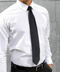 A person wearing a white PR781 dress shirt by Premier and a black tie with small white dots, standing in front of a gray concrete wall.