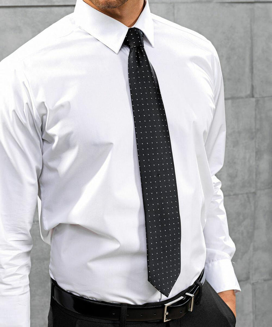 A person wearing a white PR781 dress shirt by Premier and a black tie with small white dots, standing in front of a gray concrete wall.