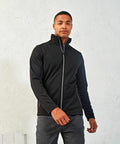 A person in a black zip-up Premier PR808 jacket poses against a light-colored, textured wall, with one hand slightly raised.
