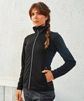 A person with a braid stands against a textured white wall, wearing a Premier PR809 and dark pants.