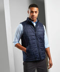 A man in a light blue shirt and a navy blue quilted Premier PR811 vest stands indoors with hands in his pockets.