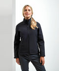 A woman with blonde hair in a ponytail is wearing a black Premier PR812 zip-up jacket and dark pants, standing indoors against a plain white background.