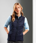 A woman with blonde hair, wearing a light blue shirt and a dark blue Premier PR815 quilted vest, stands facing forward with hands in pockets.