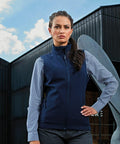 A woman stands outside in front of a building, wearing a navy Premier PR816 vest over a blue and white checkered shirt. She has long, dark hair pulled back and displays a serious expression.