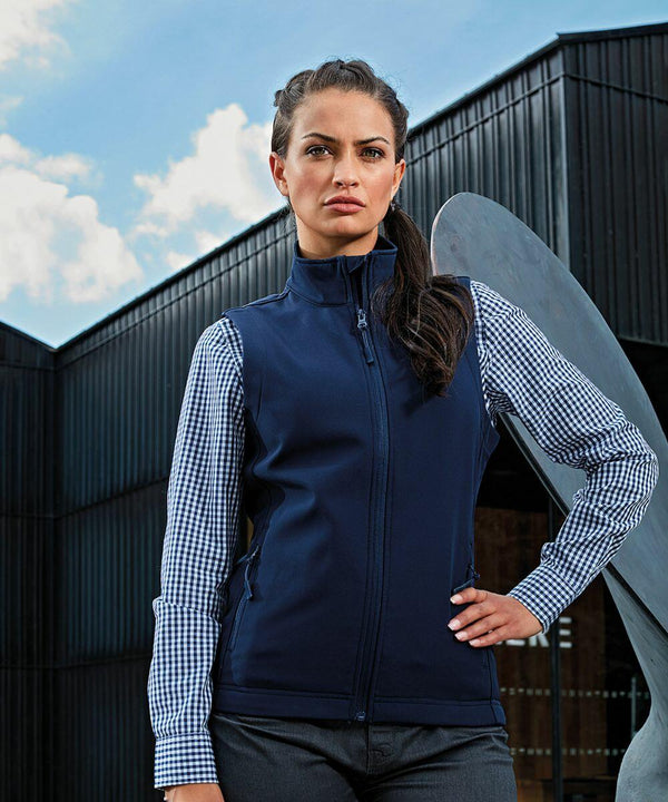 A woman stands outside in front of a building, wearing a navy Premier PR816 vest over a blue and white checkered shirt. She has long, dark hair pulled back and displays a serious expression.
