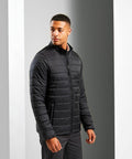 A man stands in a black, zip-up, lightweight quilted PR817 jacket by Premier and dark pants, facing slightly to the right against a plain, light gray background.