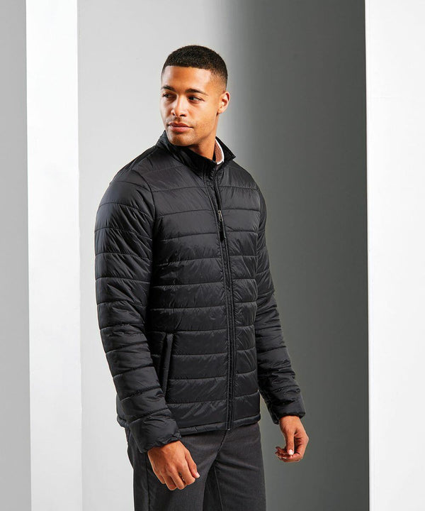 A man stands in a black, zip-up, lightweight quilted PR817 jacket by Premier and dark pants, facing slightly to the right against a plain, light gray background.