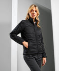 A person wearing a black Premier PR819 quilted jacket and grey pants stands with hands in pockets against a white and grey background.