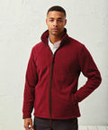 A man wearing a maroon Premier PR823 fleece jacket with hands in pockets stands against a textured white wall.