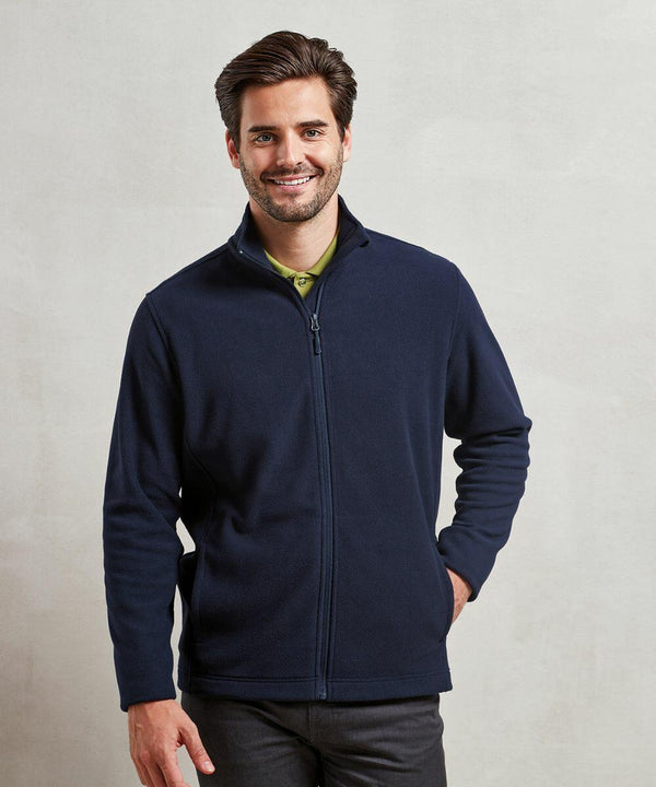A man with a beard and short hair smiles while wearing a dark Premier PR830 zip-up jacket and light green shirt. He has one hand in his pocket and stands against a plain, light-colored background.