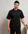 A man stands against a neutral background, wearing a short-sleeve black Premier PR900 chef's coat. He rests one hand on a wooden surface, and there are ceramic items in the background.