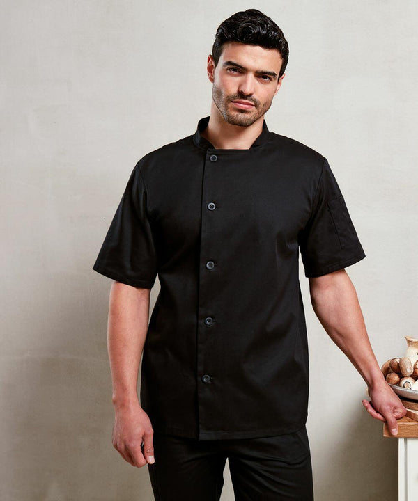 A man stands against a neutral background, wearing a short-sleeve black Premier PR900 chef's coat. He rests one hand on a wooden surface, and there are ceramic items in the background.