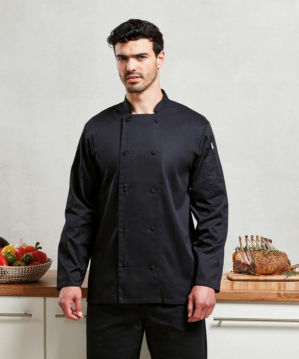 A man wearing a black chef's coat stands in a kitchen. Behind him is a counter with assorted vegetables and a carved Premier PR903 roast.