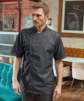 A man in a dark short-sleeved Premier PR906 chef's coat stands in a restaurant with a mix of teal, mustard, and white seating, brick walls, and framed art in the background.