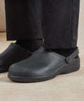 A person stands on a light-coloured floor wearing Premier Chefs non-slip Anchor clogs with a back strap, paired with black socks and black trousers.