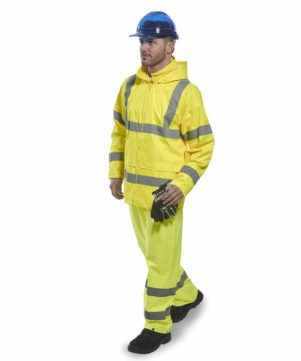 A man wears a yellow high-visibility jacket and pants with reflective stripes, a blue hard hat, and black boots, holding Portwest PW011 gloves in his right hand.
