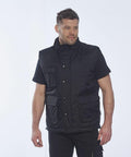 A man stands against a plain background, wearing a black short-sleeve shirt and a black Portwest PW027 utility vest with multiple pockets.