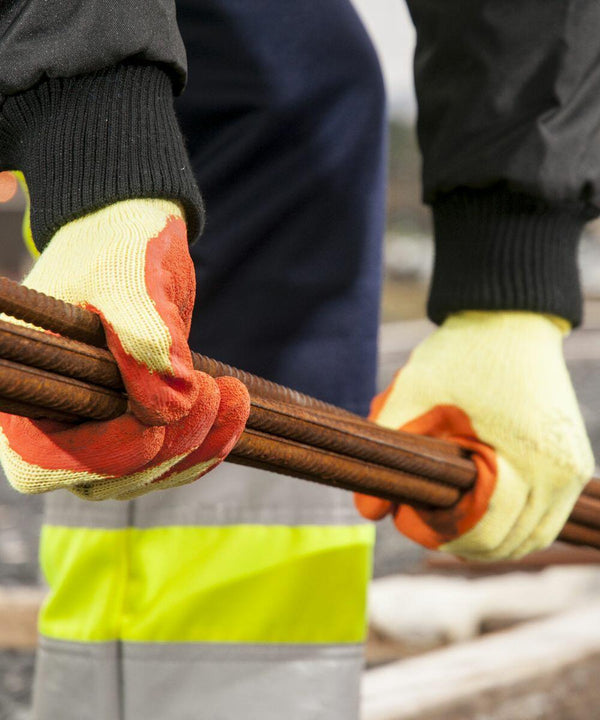 A person wearing yellow PW072 gloves from Portwest and a black jacket holds several rusted steel rods outdoors.
