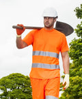 A man wearing a white hard hat, orange high-visibility shirt and pants, and gloves, holds a Portwest PW090 over his shoulder while standing outdoors.