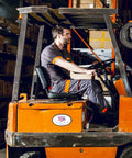 A man in a warehouse is driving an orange PW100 forklift with a "Portwest" logo on it. He is wearing a dark gray work uniform with orange accents. Shelves filled with boxes are visible in the background.