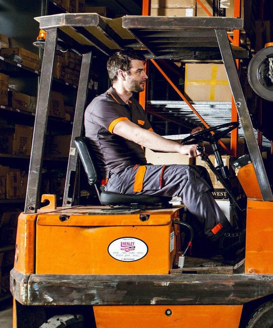 A man in a warehouse is driving an orange PW100 forklift with a "Portwest" logo on it. He is wearing a dark gray work uniform with orange accents. Shelves filled with boxes are visible in the background.