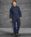 A man wearing a Portwest PW250 blue coverall stands against a gray brick wall background.