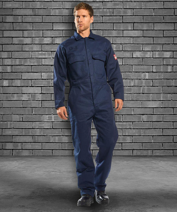 A man wearing a Portwest PW250 blue coverall stands against a gray brick wall background.