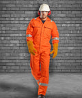 Person wearing orange PW253 safety coveralls by Portwest, yellow gloves, white hard hat, and red ear protection, walking in front of a gray brick wall.