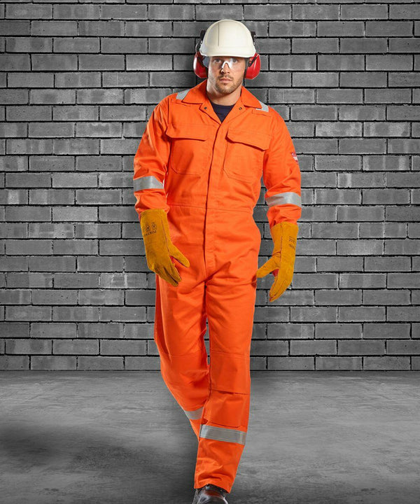 Person wearing orange PW253 safety coveralls by Portwest, yellow gloves, white hard hat, and red ear protection, walking in front of a gray brick wall.
