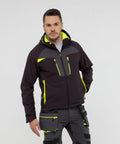 A man wearing a black and neon yellow PW263 jacket with matching pants from Portwest, standing against a plain background. The PW263 jacket has multiple zippered pockets and a hood. The pants have various pockets and straps.
