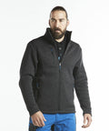 A man with a beard wearing a dark gray Portwest PW292 zip-up jacket, a black shirt, and blue pants stands upright against a white background.