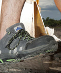 Close-up of a person's foot wearing a grey and green Portwest PW325 outdoor shoe with a rugged sole, standing on a rocky surface. A blurred background with construction materials is visible.