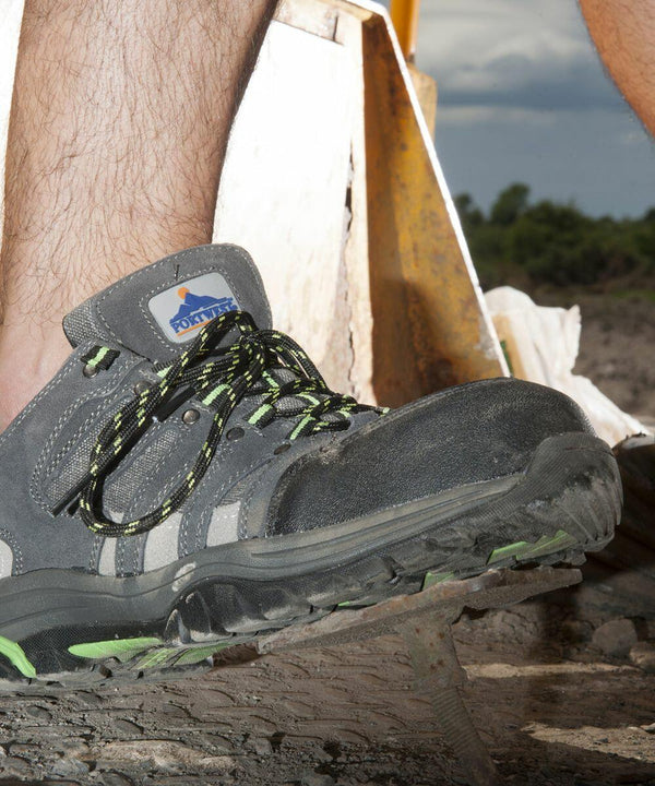Close-up of a person's foot wearing a grey and green Portwest PW325 outdoor shoe with a rugged sole, standing on a rocky surface. A blurred background with construction materials is visible.