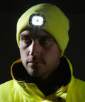 A person wearing a yellow beanie with a light attached and a yellow Portwest PW360 jacket stands against a black background.