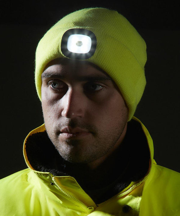 A person wearing a yellow beanie with a light attached and a yellow Portwest PW360 jacket stands against a black background.