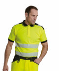 A man wearing a yellow high-visibility PW368 polo shirt with reflective stripes and black trim from Portwest stands with his hand in his pocket.