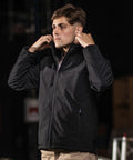 A person adjusting the collar of a zipped black PW510 jacket by Portwest, standing indoors with a dark background.