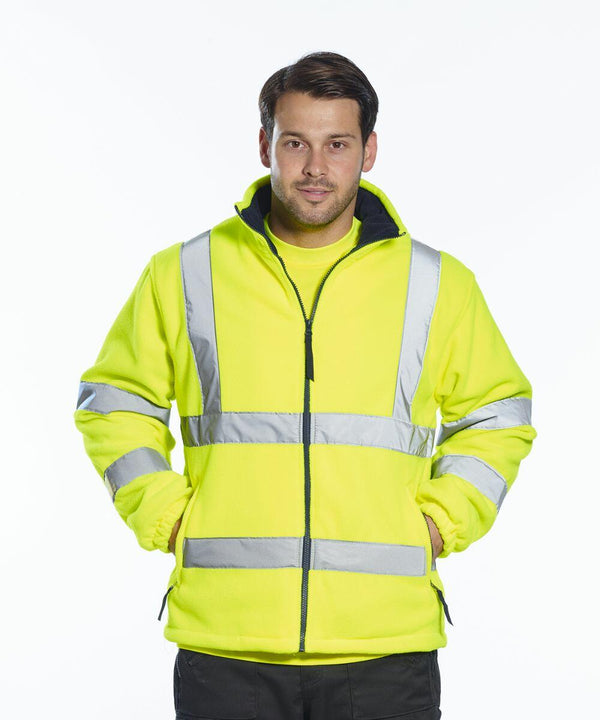 A man in a high-visibility yellow jacket with reflective stripes stands facing the camera with his hands in his pockets, wearing the Portwest PW963.