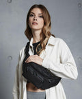 A woman with long hair wears a white jacket over a black top, holding a black crossbody bag (QD012 by Quadra). She stands in front of a gray wall.
