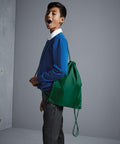 A boy wearing a blue sweater and gray pants stands with a green Quadra QD017 drawstring backpack, looking to the side with his mouth open, against a gray background.