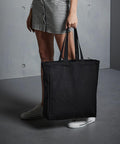 A person in checkered shorts and white sneakers holds a large Quadra QD023 tote bag, standing on a gray floor with a gray wall in the background.