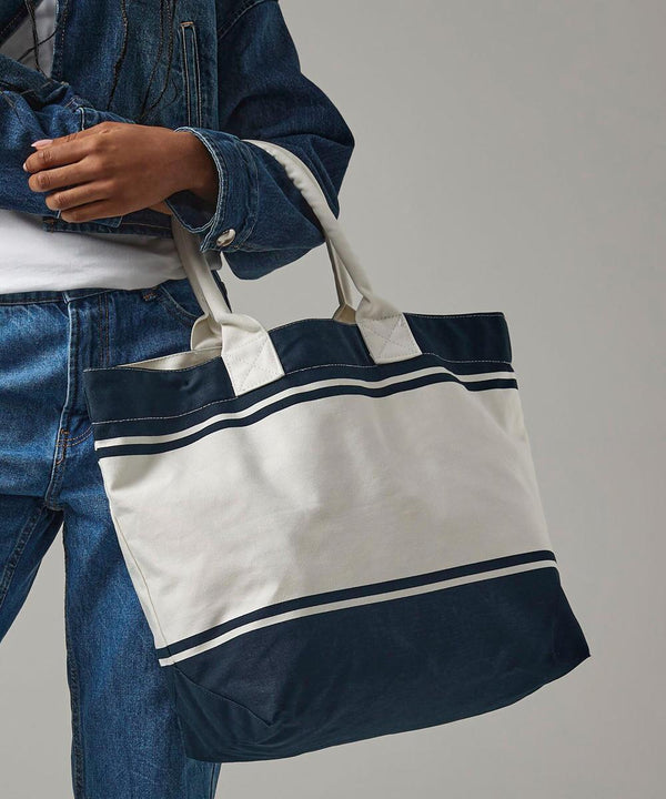 A person in denim holds the Quadra Canvas deck bag, featuring navy blue and white stripes, two handles, and a colour-blocked design. Only their torso and arm are shown.