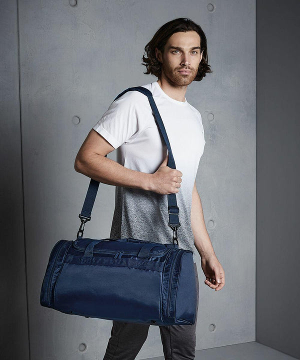 A man with shoulder-length hair, wearing a white and gray shirt, carries a dark blue Quadra QD070 over his shoulder against a gray background.