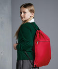 A young girl with long hair, wearing a green sweater and a gray skirt, stands with a red Quadra QD071 drawstring backpack.