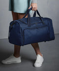 Person holding a large blue Quadra QD074 duffel bag, standing on a gray surface while wearing white sneakers and light blue clothing.