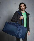 A person with shoulder-length hair holds a large blue Quadra QD080, wears a green shirt under a black jacket, and stands against a gray, textured background.