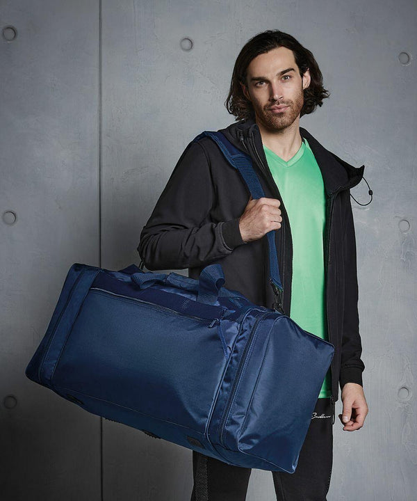 A person with shoulder-length hair holds a large blue Quadra QD080, wears a green shirt under a black jacket, and stands against a gray, textured background.