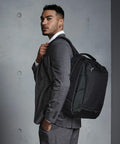 A man in a gray suit carries a black Quadra QD269 backpack over his shoulder, looking to his left. The background is a gray wall with round button-like indents.