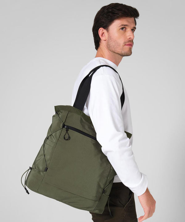 A man in a white long-sleeve shirt carries the Quadra UrbanShift tote, a large olive green bag with black straps and a front zipped pocket, over his shoulder against a plain light grey background.