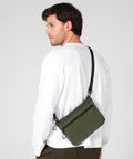 A man with short brown hair wears a white long-sleeve shirt, dark trousers, and carries the Quadra UrbanShift sacoche—an olive green crossbody bag with a black zip and strap. The background is plain light grey.